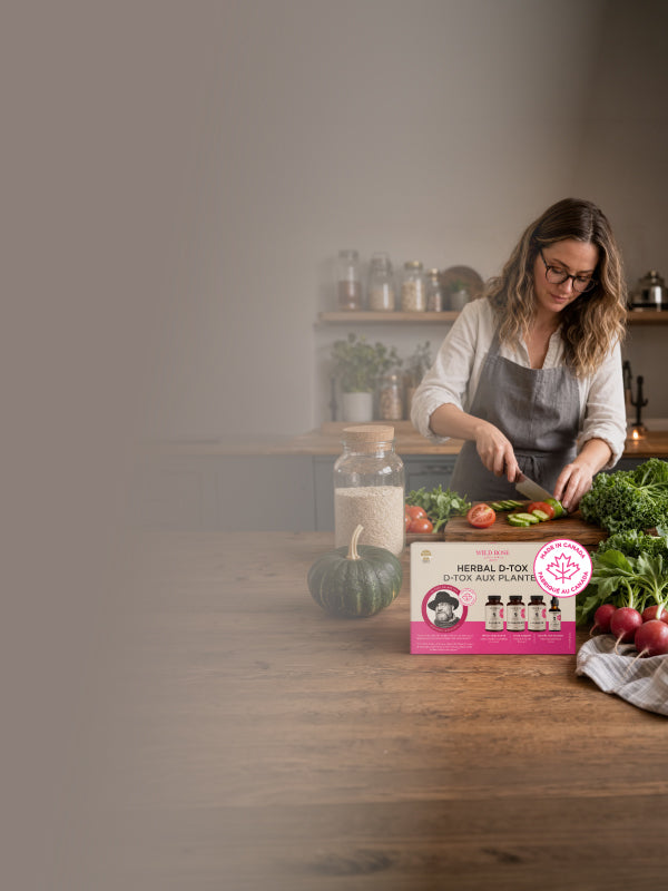 A woman in a kitchen wearing an apron and glasses, preparing food at a wooden table. In the foreground, a pink box labeled 'Herbal Super Quick and Plant' with small bottle illustrations is placed next to a green pumpkin, a glass jar, and fresh vegetables.